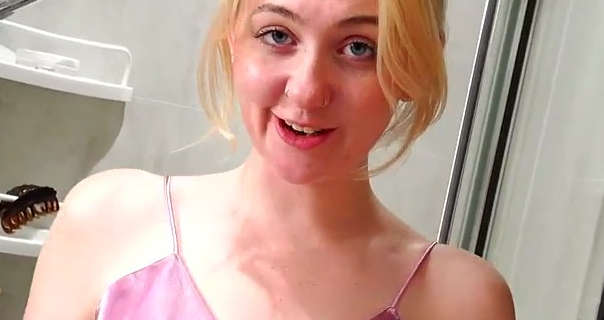 Em Yang smirks and looks into the camera. She wears her long blonde hair tied back and up in a bun. Em stands in front of a wall of cream bathroom tiles with a chrome shower area and small white shelves to the side. She appears in a light pink nightie, soft foundation, mascara and a pink lipstick.