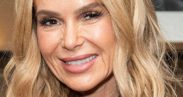 Amanda Holden smiles as she stands in front of a wooden display area. She has her blonde hair loose and in waves. Amanda is seen in black mascara, dark pink lipstick and a matt foundation.