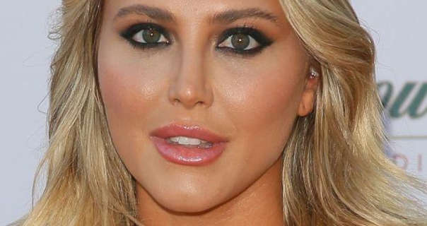 Cassie Scerbo is seen with her long blonde hair in soft waves and loose around her shoulders. She wears a matt foundation with pink blush, black mascara, eyeliner and a pink gloss lipstick. Cassie stands in front of a dark white wall which has green, gold and red text on it.