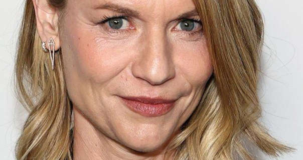 Claire Danes is seen in a soft blush, mascara, red lipstick and two silver earrings. She stands in front of a dark white wall. Claire has her shoulder length blonde hair styled in waves.