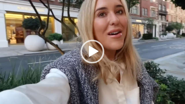 Em Sheldon appears with her long blonde hair loose and down. She sits on a low wall surrounded by green shrubs on a central London street. She wears a grey mix knitted jump across her shoulders. She stars in a cream shirt dress, matt foundation, mascara and a gloss baby pink lipstick.