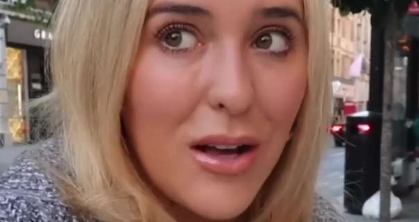 Em Sheldon glance to one side as she talks. She wears her long thick blond hair loose and straightened. Em is seen in a matt foundation, blush, mascara and a pink gloss lipstick. She wears a grey mix jumper across her shoulders as she sits outside in central London.