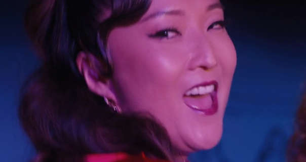 Ashley Park is seen in front of a blue and black screen as she performs on stage. Ashley has her thick hair pinned back and in waves. She wears a matt foundation with mascara and a dark pink lipstick.