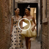 Lily Collins and Ashley Park hold hands and walk down a narrow cobblestone alleyway in Venice. Lily wears a black and white spotty top and skirt and Ashley a yellow puff dress. They both have their hair loose and tied back with natural makeup and gold jewellery.