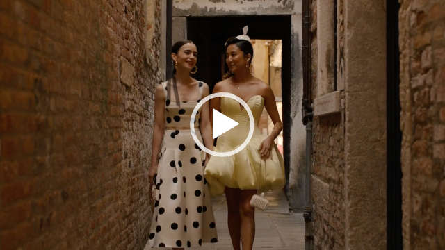 Lily Collins and Ashley Park hold hands and walk down a narrow cobblestone alleyway in Venice. Lily wears a black and white spotty top and skirt and Ashley a yellow puff dress. They both have their hair loose and tied back with natural makeup and gold jewellery.