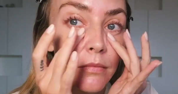 Janni Delér rubs in concealer with both of her hands. She sits in front of a white block display wall. Janni wears her long dark blonde hair pushed back. She is seen in a light cream dressing gown.