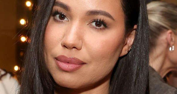 Jurnee Smollett stands in a small room lit by several orange light bulbs. She appears with her long black hair loose and straight. Jurnee wears light foundation with a little blush, mascara and a mauve lipstick. There are other guests at the nighttime event around her.