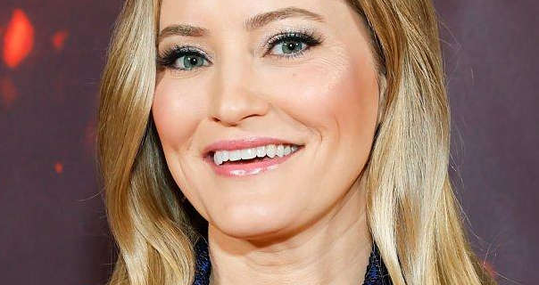 Justine Ezarik appears in front of a brown wall that has a few red flecks on it. She wears her long blonde hair loose and in soft waves. Justine is seen in a matt foundation, blush, thick black mascara and a light pink gloss lipstick.