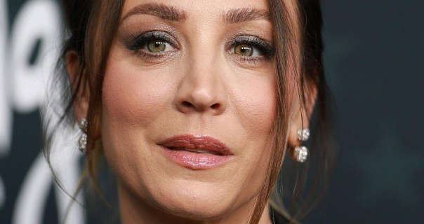 Kaley Cuoco appears in front of a dark grey wall that has white handwritten text on it. She is seen with her long dark brown hair tied back and with subtle highlights. Kaley wears a matt foundation with light pink blush, black mascara and a pink gloss lipstick with a pair of hanging stud diamond earrings.