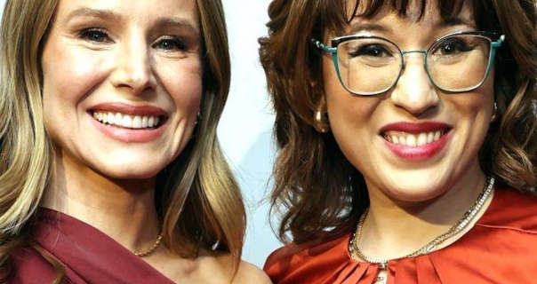 Kristen Bell stands alongside Dani Bowman. They are seen in front of a white wall. Kristen has her long blonde hair loose and a little curly. She wears a one shoulder maroon with a thin necklace, matt foundation, mascara and a pink sheen lipstick.