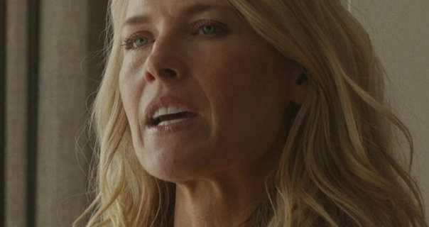 Ali Larter is seen in front of a dark beige wall and curtains. She stands with her long blonde hair loose and curled in waves. Ali wears a light foundation with lipstick and black mascara.