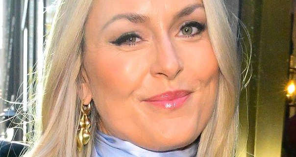 Lindsey Vonn is seen outdoors in the sunshine on a New York City street. Her long blonde hair is loose and straightened. Lindsay wears a light blue high neck top with a pair of dangling gold earrings. She appears in a matt foundation, light pink lipstick, mascara and a pink blush.