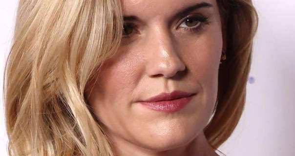 Maggie Grace is seen standing in front of a dark white event wall. She wears her thick blonde hair loose and in soft waves. Maggie appears in a dewy foundation, pink blush, mascara and a dark pink lipstick sheen.