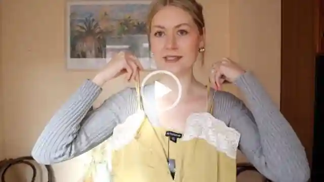 Meg Says wears a thin ribbed light grey top with her blonde hair tied back in a bun. She stands in front of pastel orange wall in her new house. Meg holds up an olive green satin and white lace camisole. She is seen in a matt foundation, pink lipstick and a pair of large gold earrings.