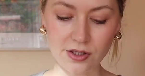 Meg Says stands in front of a light pastel orange wall in her kitchen. Meg has her long blonde hair tied up and back in a bun. She is seen in a matt foundation, blush, mascara and a dark pink lipstick. Meg wears a thin grey long sleeve top with a pair of chunky gold earrings.