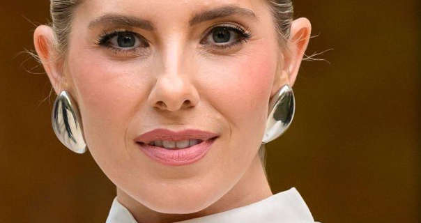 Mollie King wears her blonde hair tied up and back in a high bun. She is seen in a pair of light gold large earrings and a high neck white top. Mollie appears in a matt foundation, pink blush, mascara and a baby pink lipstick.