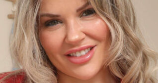 Siobhan Graves is seen in a beige wall bedroom. She has her thick long blonde hair styled in waves. Siobhan wears a red blouse with a matt foundation, mascara and a pink gloss lipstick.