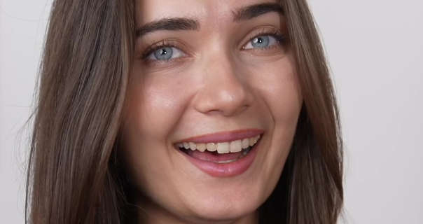 Roxxsaurus laughs and looks into the camera. She has her long light brown hair loose and straight. Roxi is seen in front of an off-white wall. She wears a light foundation with natural lashes and a dark pink lipstick.