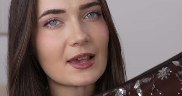 Roxxsaurus is seen with her long brown hair straightened and loose. She sits in front of a dark white painted wall. Roxi holds up a brown and silver Shein dress. She appears in a matt foundation, mascara, blush and a dark red lipstick.