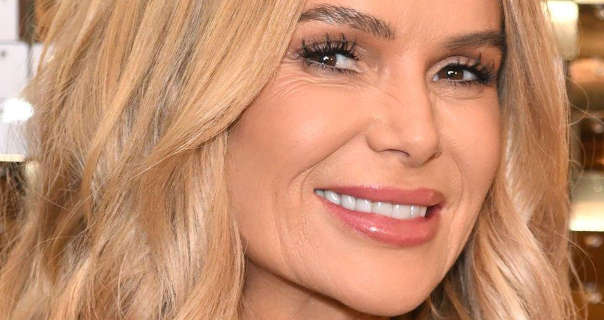 Amanda Holden wears her long blonde hair loose and in soft waves. She stands in front of a shimmering gold wall. Amanda is seen in a matt foundation, heavy mascara and a pink gloss lipstick.