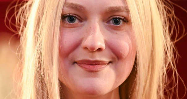 Dakota Fanning is seen against a dark red and light orange backdrop. Her long blonde hair is loose, straightened and a little frizzy. Dakota is seen in a soft foundation, mascara and a dark pink lipstick.