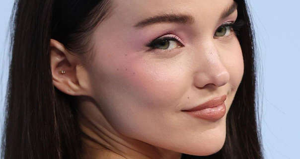 Dove Cameron wears a matt foundation with mascara, blush, dark pink lipstick and an inner ear stud earrings. She appears in front of a dusky blue wall. Doe has her long black hair down and straightened.