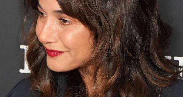 Emmanuelle Chriqui is seen turning to one side. she has her hair loose and in curls. Emmanuelle stands in front of a dark grey poster wall with white typeface writing on it. She wears a dark red lip stain with foundation and a little mascara.
