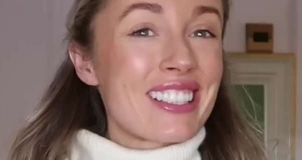 Josie Irons smiles widely showing her teeth. She stands in a powder pink room in her countryside home. Josie has her long blonde hair pinned back. She wears a soft blush with mascara, pink gloss lipstick and a high neck white top.