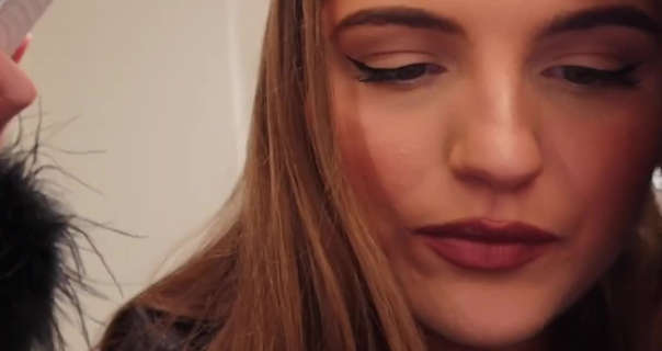 Grace Foley leans into the camera looking down towards her phone. She wears a black pyjama top in a white wall room. Grace has a matt foundation with a little blush, winged eyeliner and a dark lipstick. She is seen with her brown hair down and straightened ready for styling.