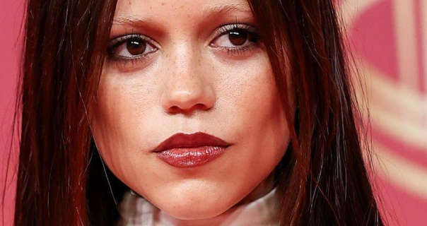 Jenna Ortega stands in front of a pastel red and light gold event wall. She has her black hair straightened, loose and with red tinting. Jenna is seen in a very light foundation, black mascara and a deep red gloss lipstick.