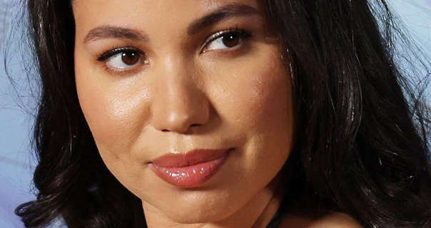 Jurnee Smollett looks to one side while standing in front of a light ice blue poster wall. She has her long thick black hair loose and naturally curled. Jurnee wears a soft blush with light foundation, mascara and a dark pink gloss lipstick.