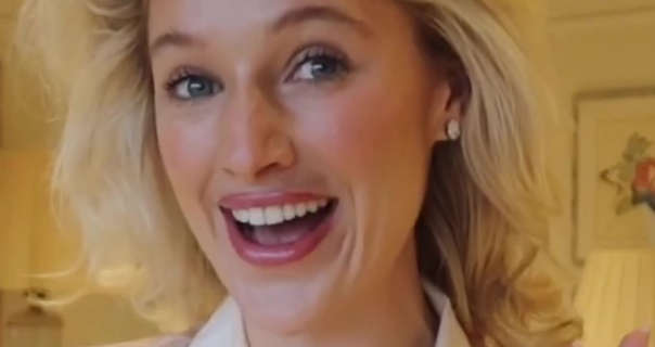 Leonora Smee talks to the camera as she gets ready. She stands in her large filming bedroom at home. Leonora has her hair styled in heatless curls. She wears a heavy foundation with blush, mascara, large gemstone earrings and a dark pink gloss lipstick.