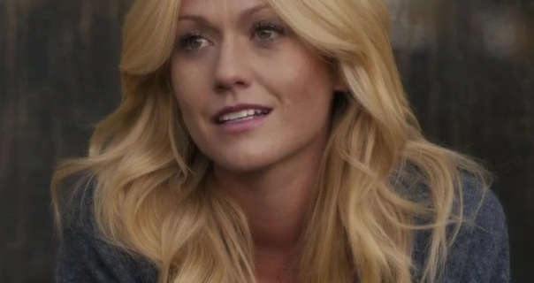 Katherine McNamara stands outside on the deck of a wooden countryside cabin. Her long blonde hair is down, loose and volumised. Katherine wears a thin grey cardigan with a matt foundation, mascara and a dark pink lipstick.