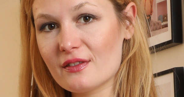 Freya H stands on the Only Tease staircase in front of a wall of framed model images. Freya has her blonde hair loose and tucket behind one ear. She wears a light founbdation with blush, natural eyelashes and a pink gloss lined lipstick.