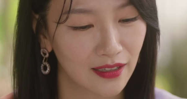 Baek Eun-hye is seen in a leafy restaurant dining area during the day. She has her long black hair straightened and loose. Baek Eun-hye wears a matt foundation with blush, red lipstick and a pair of silver two circle hanging earrings.