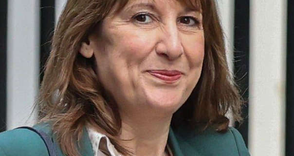 Rachel Reeves walks past a row of high black metal railings and a white building wall. She has her light brown hair loose and styled naturally. Rachel is seen in a dark teal blazer and gold shirt. She wears blush with foundation and a dark pink lipstick.