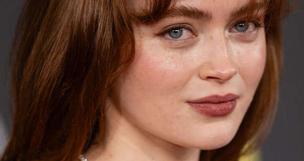 Sadie Sink wears her hair loose, pushed back and with a long fringe. She stands in front of a grey and yellow event wall. Sadie is seen in a light foundation, natural eyelashes and a dark pink lipstick.