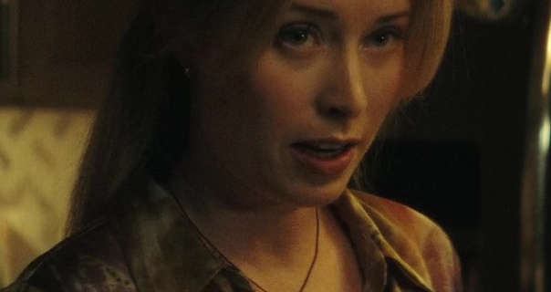 Hannah Emily Anderson stands in a dark kitchen in front of a wall of wood cabinets. She wears her hair loose and pinned back. Emily is seen in soft foundation, dark pink lipstick and a long sleeve shirt.