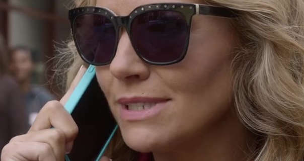 Becki Newton is seen holding a phone in a bright blue case to an ear. She has her long blonde hair loose and in waves. Becki appears in a pair of large black frame sunglasses. She wears a matt foundation with a light pink lipstick.