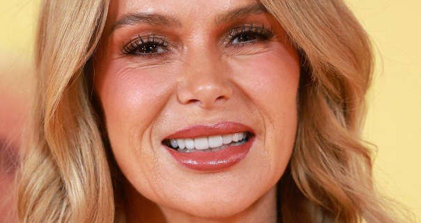 Amanda Holden appears in front of a pale yellow and light brown film poster wall. She has her long blonde hair loose and in soft curls. Amanda wears blush with black mascara and a dark red gloss lipstick.