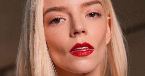 Anya Taylor-Joy tilts her head a little to the side and gazes into the camera. She stands in front of a large open staircase at night. Anya has her long blonde hair straightened and loose. She is seen in a contouring foundation, mascara and a dark red lipstick.