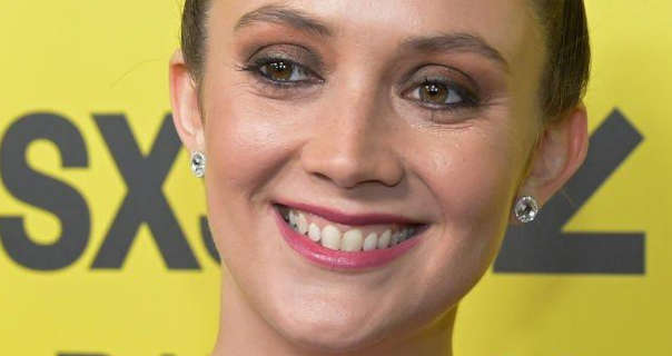 Billie Lourd smiles as she looks to one side. She stands in front of a yellow wall with light grey text on it. Billie has her brown hair tied up in a bun. She appears in a soft foundation, mascara, dark pink lipstick and a pair of large diamond stud earrings.