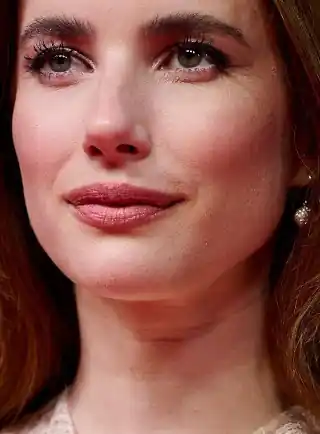 Emma Roberts looks into the distances with a small smile. She is seen with her long red tinted hair loose and straightened. Emma wears a full foundation with globe earrings, sheen lipstick, soft blush and black mascara.