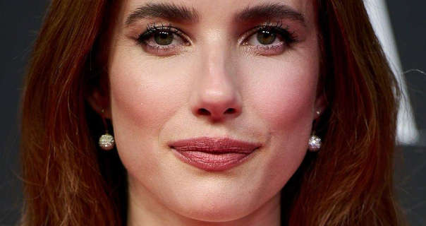 Emma Roberts stands in front of a dark grey event wall that has large white text on it. She wears her long hair loose and tinted with red. Emma stars in a matt foundation, mascara, dark pink sheen lipstick and a pair of hanging gemstone sphere earrings.