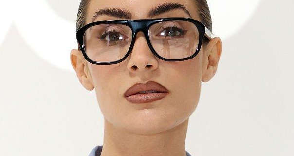 Frankie Bridge is seen with her hair tied back in a slick bun. She wears a pair of large black frame glasses with a smoothing foundation, blush, mascara and a dark pink lipstick. Frankie stands in front of a cream and white studio wall.