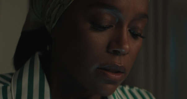 Aja Naomi King sits in a dark living room with her eyes partially closed. She wears her black hair tied back and covered with a sleep scarf. Aja stars in a long sleeve white and green stripe pyjama top.
