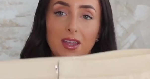 Hazel Maria Wood sits in front of a large cream abstract painting. She wears her long hair straightened and loose. Hazel appears in heavy foundation, metal earrings, eye liner and a gloss lipstick. She holds a pair of Zara shorts up to the camera.