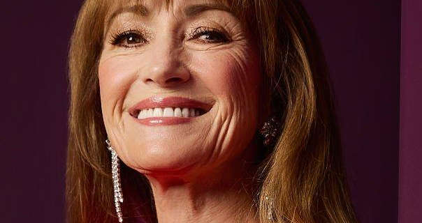 Jane Seymour is seen in front of a light and dark maroon wall. She has her long hair loose and in soft waves. Jane wears blush with mascara, dark pink lipstick and a pair of hanging gemstone earrings.