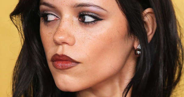 Jenna Ortega appears with her thick black hair loose and slightly wavy. She wears a soft blush with black eyeliner, mascara and a dark red overlined lipstick. Jenna glance to the side and turns her head. She is seen in front of a dark yellow wall in a pair of silver globe earrings.