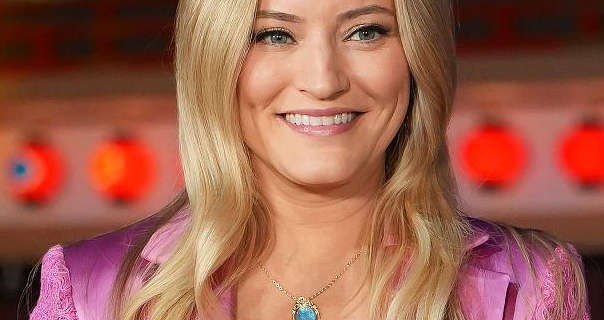 Justine Ezarik smiles and looks to the side. She stands in front of a row of red lights at an event outdoors at night. Justine has her long blonde hair loose and curled at the ends. She wears a pink blazer with a matt foundation, black mascara, dark eye shadow and a pink lipstick.